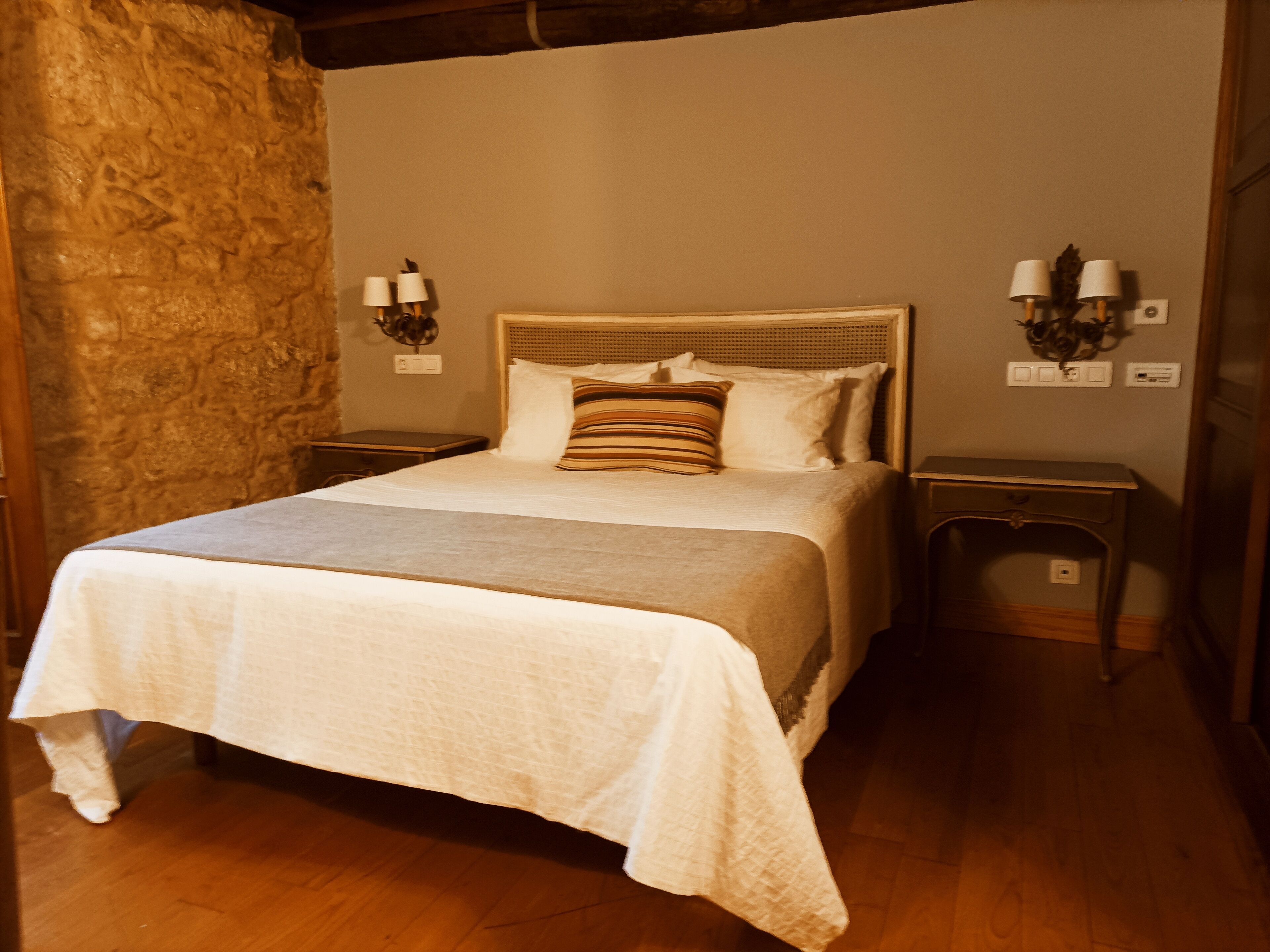 A neatly made bed with a striped pillow, two bedside lamps, and a stone wall to one side.