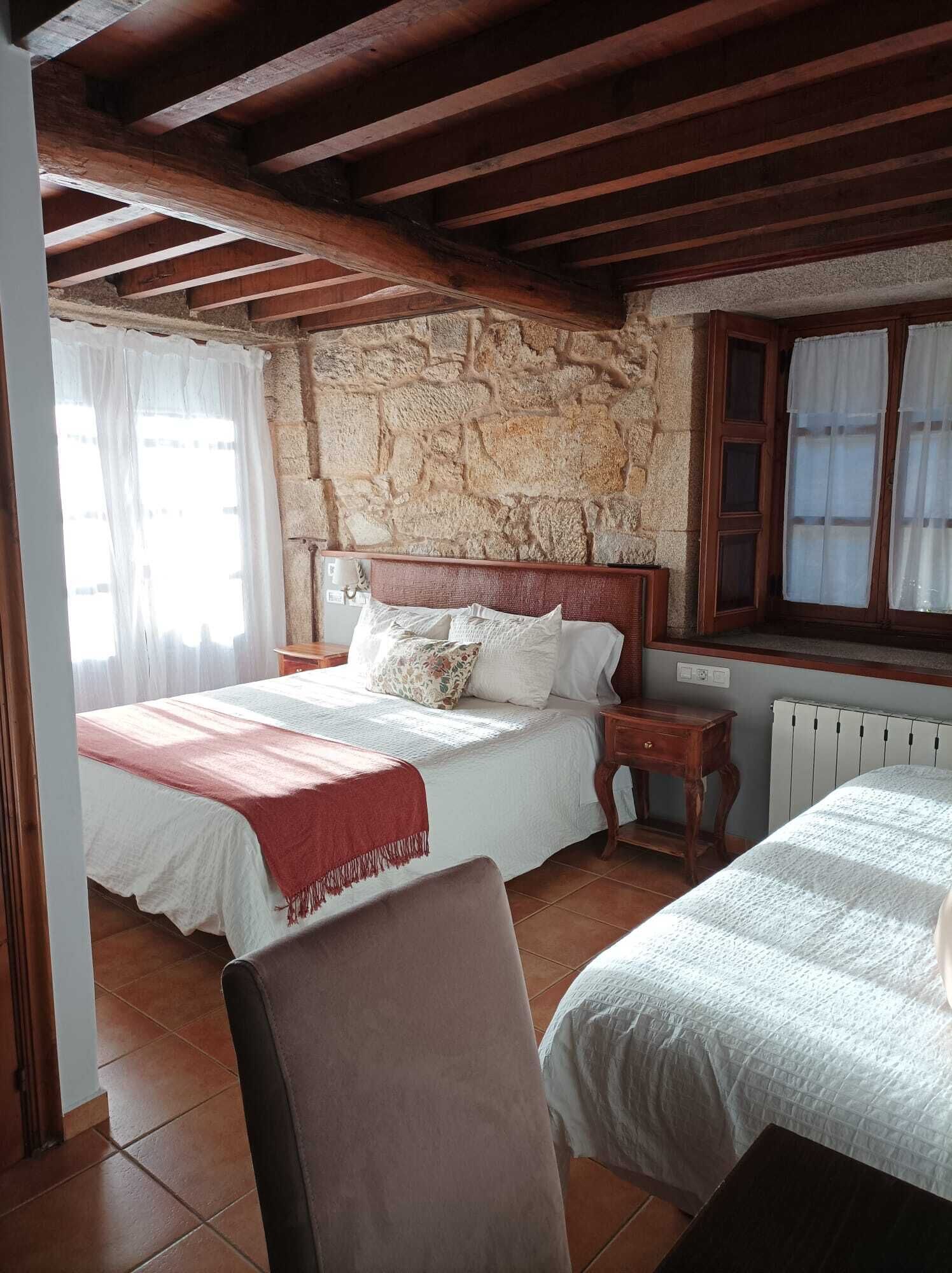 Un dormitorio con pared de piedra, vigas de madera, una cama con ropa blanca, una silla y una ventana con cortinas.