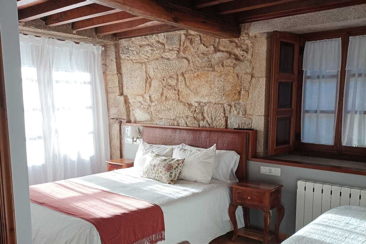 A bedroom with a stone wall, wooden beams, a bed with white bedding, a chair, and a window with curtains.