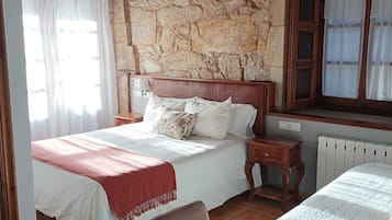 A bedroom with a stone wall, wooden beams, a bed with white bedding, a chair, and a window with curtains.