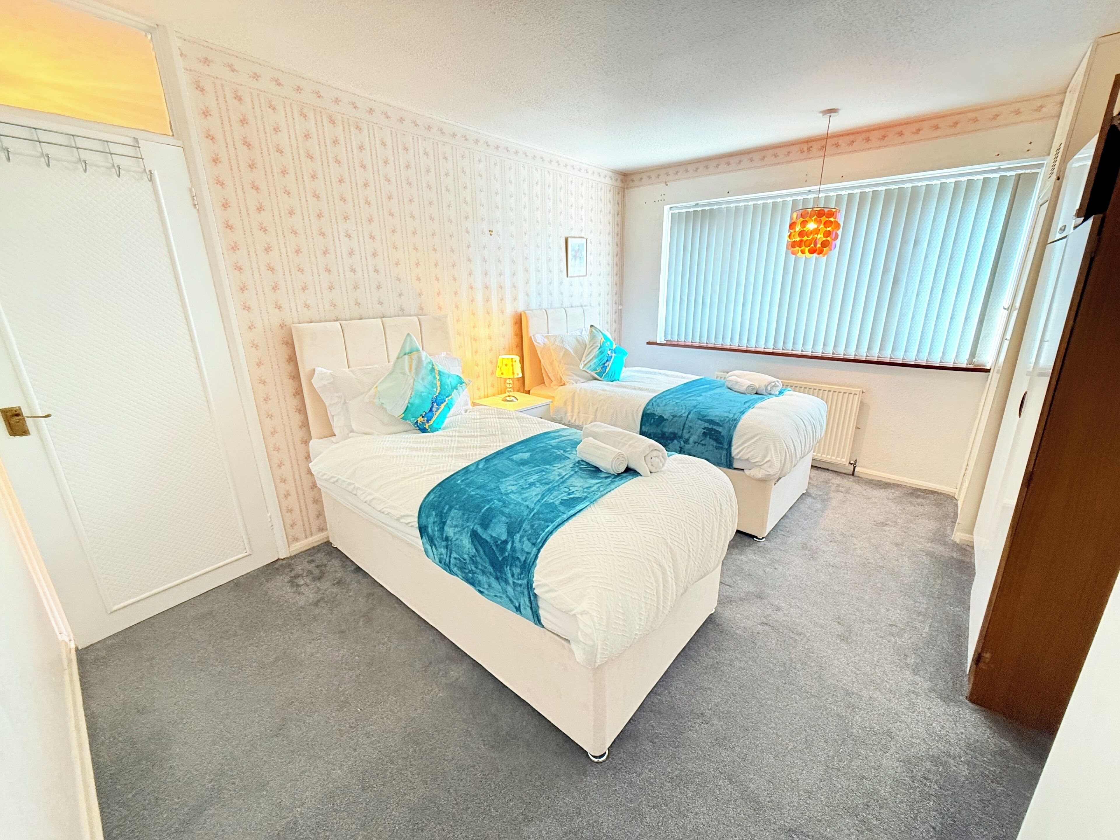 A hotel room with two beds, a window with blinds, a radiator, and a small table with a lamp.