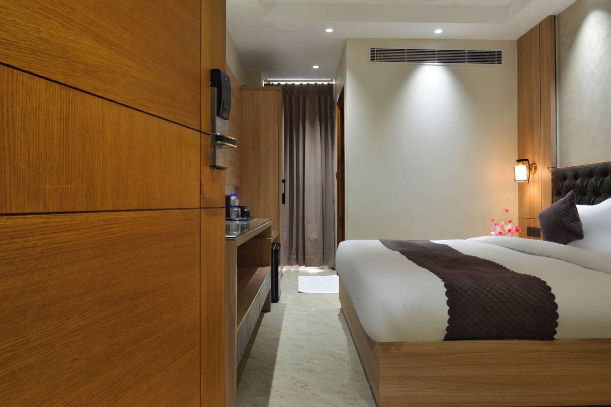 A hotel room with a wooden wardrobe, a bed with a dark quilt, a bedside table with a lamp, and a window with curtains.