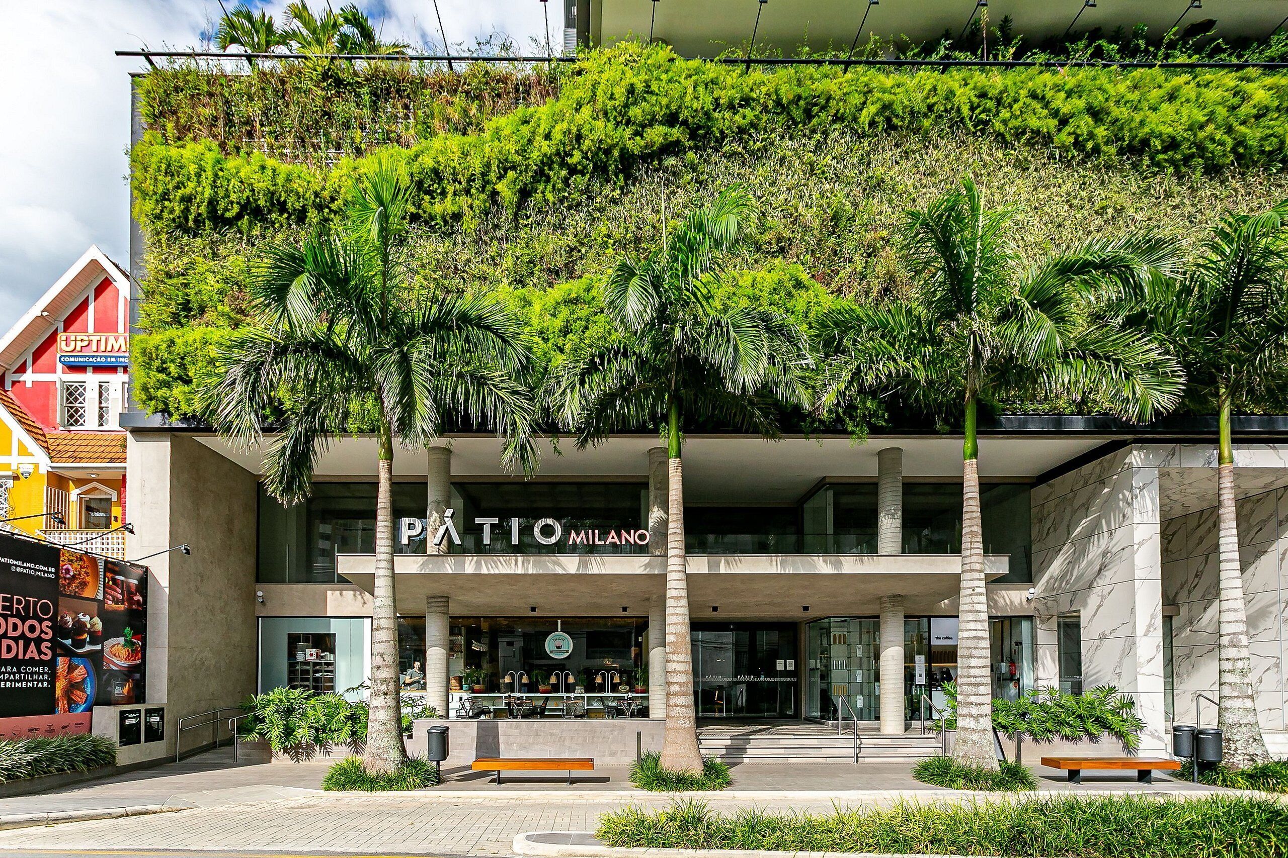 Ein modernes Gebäude mit vertikaler Begrünung, Palmen und einem Schild mit der Aufschrift 'PATIO MILANO'.