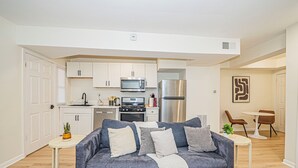 Ein kompakter Wohnbereich mit Küchenzeile, grauem Sofa und einer Essecke mit modernen Stühlen und einem kleinen Tisch.