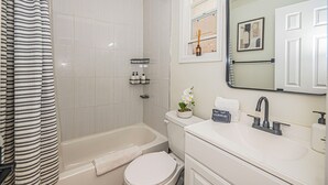 Ein modernes Badezimmer mit Badewanne, Toilette und Waschbecken. Ein Duschvorhang, ein Handtuch und ein Spiegel über dem Waschbecken sind vorhanden.