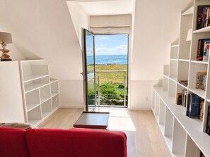Une piĂšce avec un canapĂ© rouge, une bibliothĂšque et une vue sur lâocĂ©an, visible Ă travers une porte vitrĂ©e.