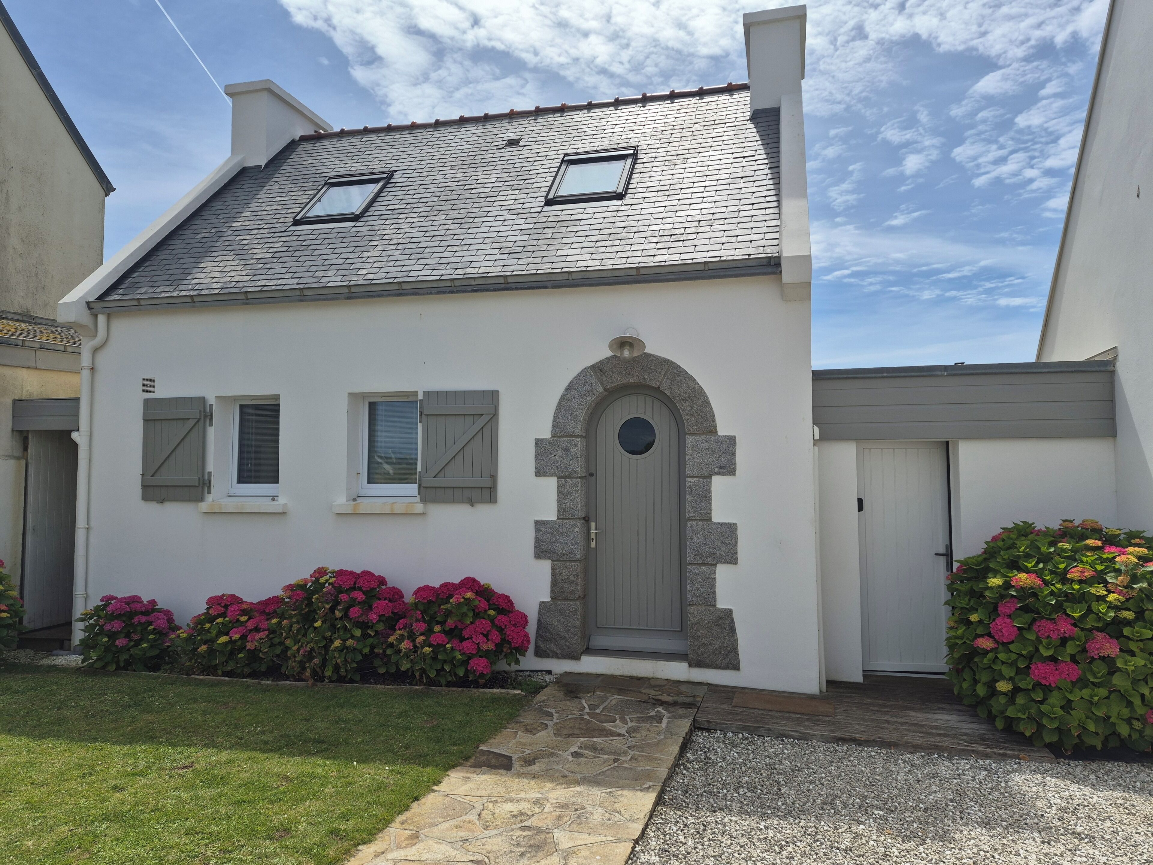 Une maison à deux étages, avec un toit recouvert de tuiles grises, une porte centrale surmontée d’un arc en pierre et une petite fenêtre à l’étage supérieur.