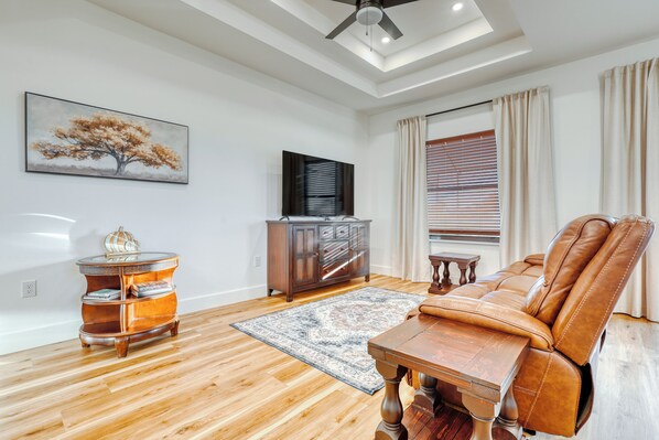 A living room with a wooden floor, a brown leather sofa, a wooden coffee table, a TV on a cabinet, and a large window with blinds. - Waterview Villa With Heated Pool (Rotonda West)