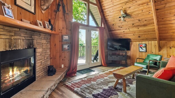 Interior de una cabaña de troncos con chimenea de piedra, paredes de madera, un sofá de cuero verde y un televisor.