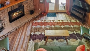Interior acogedor de una cabaña con chimenea de piedra, ventilador de techo y un amplio ventanal con vistas al exterior.