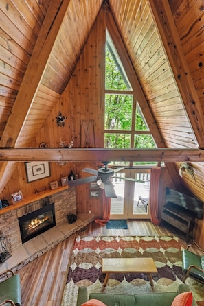 Ein gemütliches Blockhäuschen-Interieur mit Kamin aus Stein, Deckenventilator und großem Fenster mit Blick ins Freie.