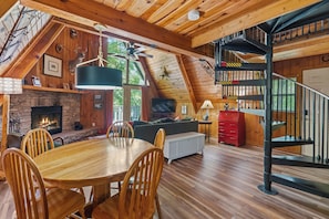 Ein gemütliches Blockhaus-Interieur mit Essbereich, Kamin, Holzbalkendecke und einer Wendeltreppe.