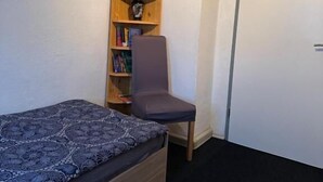 A small room with a single bed, a bookshelf, a chair, and a door. - Corner Haus RM2 (Schnaittach)