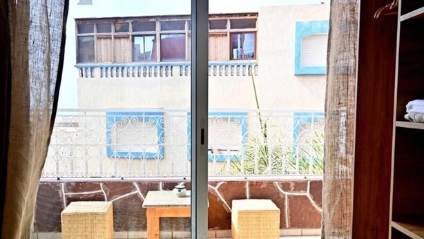 A balcony with a table and two wicker stools, a metal railing, and a view of a building with blue trim and a window. - Standard Double Room #1 (Tamraght Ouzdar)