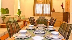 A dining area with a round table set for six, surrounded by wicker chairs, with a sofa and a TV in the background.