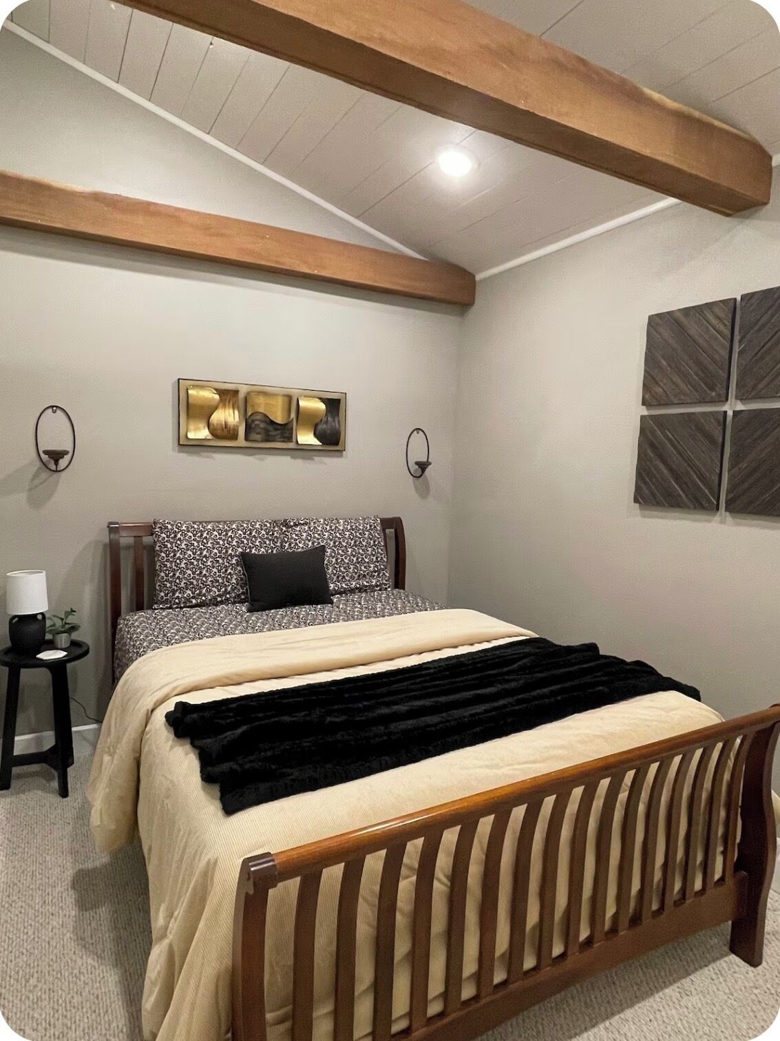 A bedroom with a wooden bed frame, a headboard, a bedside table with a lamp, and a wall-mounted artwork.