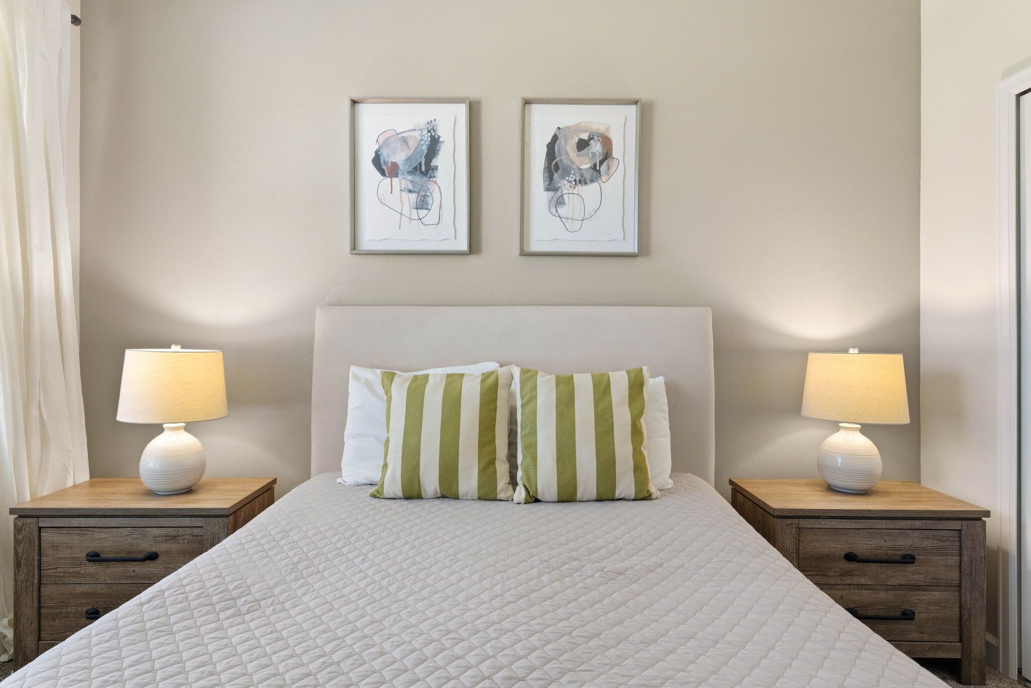 Una cama bien hecha con almohadillas a rayas, dos mesitas de noche con lámparas, y dos cuadros abstractos en la pared.