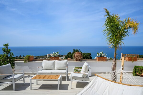 A terrace with white furniture, a hammock, and potted plants against a backdrop of the sea.