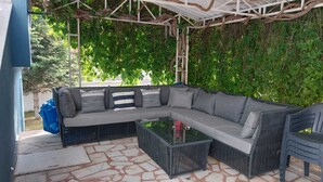 Un espace extérieur aménagé, abrité et doté d’un canapé d’angle, d’une table basse en verre et d’un fauteuil en osier.