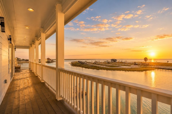 A covered porch with a view of a sunset over water, wooden flooring, and a railing.