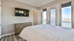 Ein Schlafzimmer mit einem großen Bett, einem Fernseher an der Wand, einem Schminktisch und einem Balkon mit Blick auf den Strand.