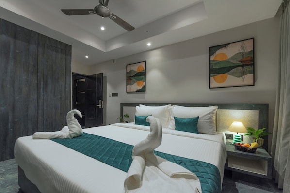 Una habitación de hotel con una cama grande, dos cuadros colgados en la pared, un ventilador de techo y una mesita de noche con lámpara y frutero.