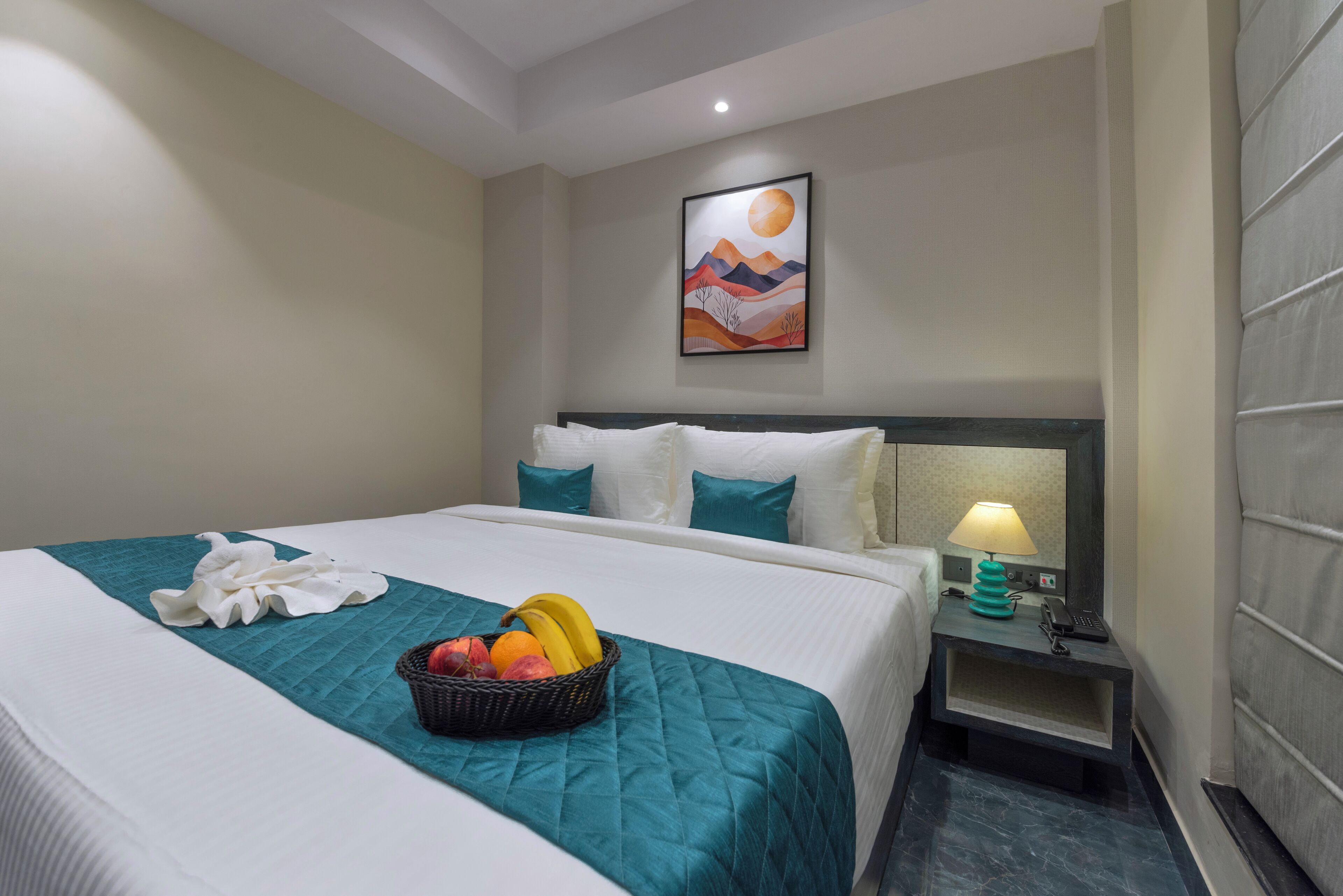 Une chambre d’hôtel avec un lit, une table de chevet avec une lampe, un tableau accroché au mur et un panier de fruits posé sur le lit.