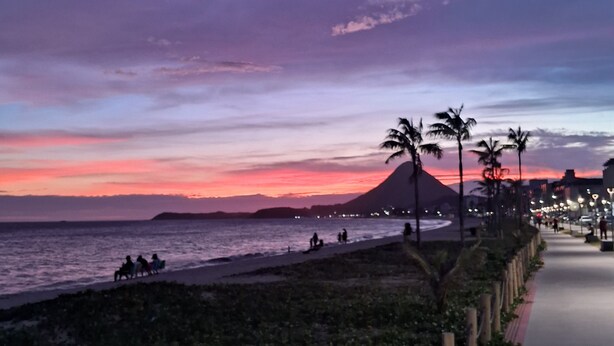 Une vue sur le coucher de soleil sur la côte, avec une promenade dallée, des palmiers et une montagne lointaine.