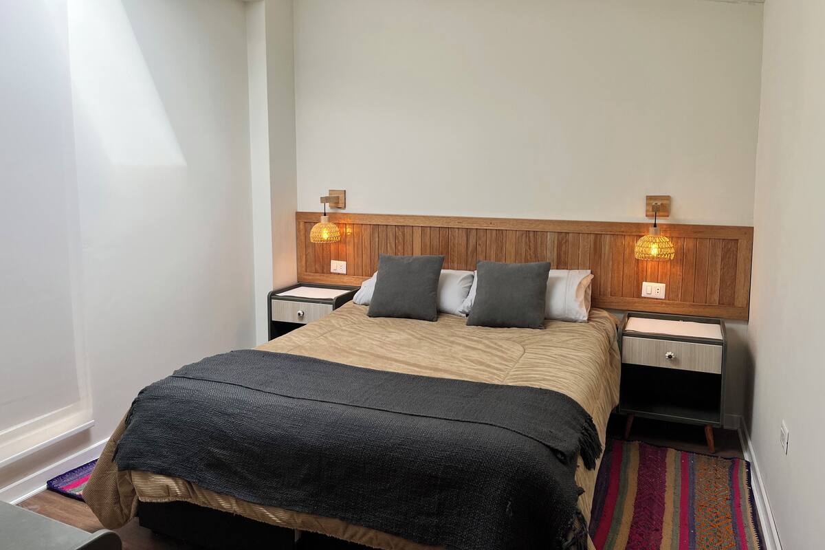Quarto com cabeceira de madeira, duas luminárias de apoio, cama com manta escura e tapete colorido.