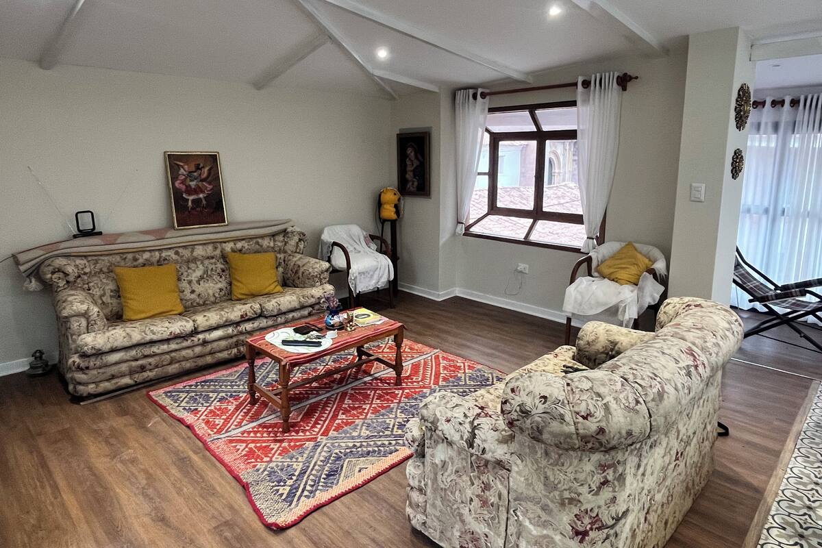 Sala de estar com sofá, poltrona e mesa de centro. Há uma janela com cortinas e vista para o exterior.