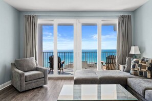 Een ruime woonkamer met een grote schuifdeur naar een balkon met uitzicht op de oceaan. Er zijn twee fauteuils en een bank, een salontafel en een lamp op een bijzettafeltje.