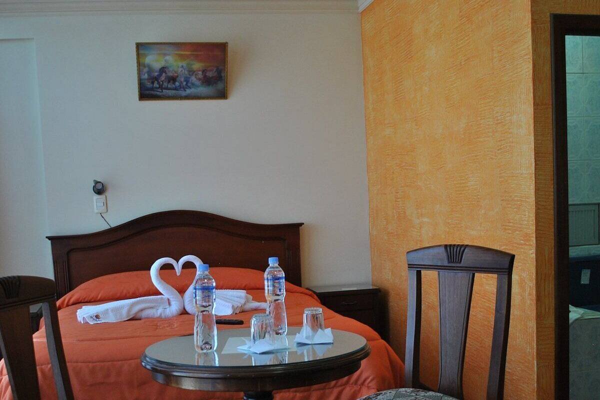 Une chambre d’hôtel avec un lit, deux chaises, une table ronde avec des bouteilles d’eau et un tableau accroché au mur.