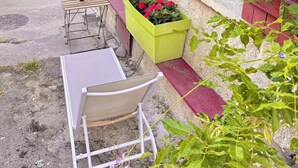 Uma viela estreita com uma mesa e banco de madeira, um canteiro verde com flores vermelhas, e um edifício com portas azuis e uma cerca vermelha.