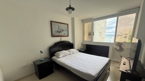 Een slaapkamer met een bed, nachtkastje, bureau, stoel, raam met uitzicht op gebouwen en een plafondventilator.