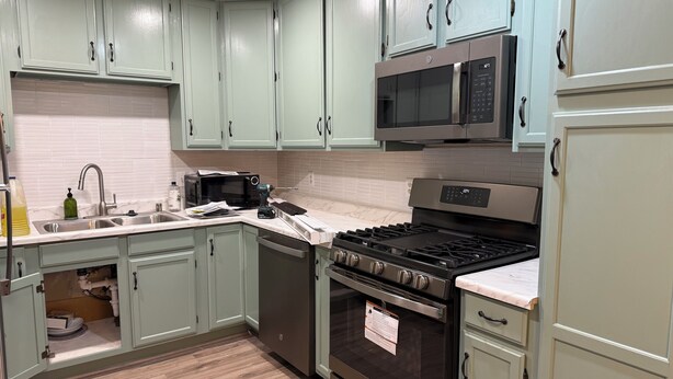 Una cocina con gabinetes verde claro, un lavamanos de acero inoxidable, un microondas, una estufa y un lavavajillas.