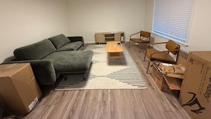 Una sala de estar con un sofá verde, una mesa de centro de madera y dos sillas. Hay cajas en el suelo, lo que indica una mudanza reciente.