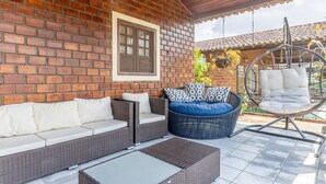Une terrasse couverte, meublée de meubles en osier, avec un fauteuil suspendu et un mur en briques.