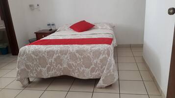 Ein Bett mit floralem Überwurf, einem roten Kissen und einem roten Akzent auf dem Überwurf. Ein Nachttisch mit zwei Wasserflaschen und einer Fernbedienung steht daneben. Der Raum hat weiße Wände und Fliesenboden.