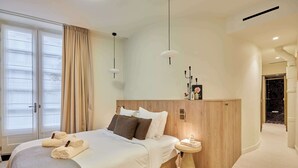 Habitación de hotel moderna con una cama grande, una mesita de noche, una pequeña mesa redonda y vistas al exterior a través de una ventana con cortinas.