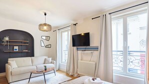 Ein modernes Hotelzimmer mit Sofa, kleinem Couchtisch, Bett, Fernseher und großen Fenstern mit Vorhängen.