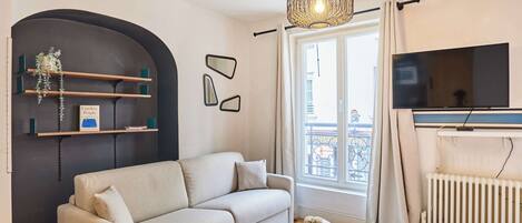 A living room with a sofa, a coffee table, a TV, a radiator, and a window with curtains.