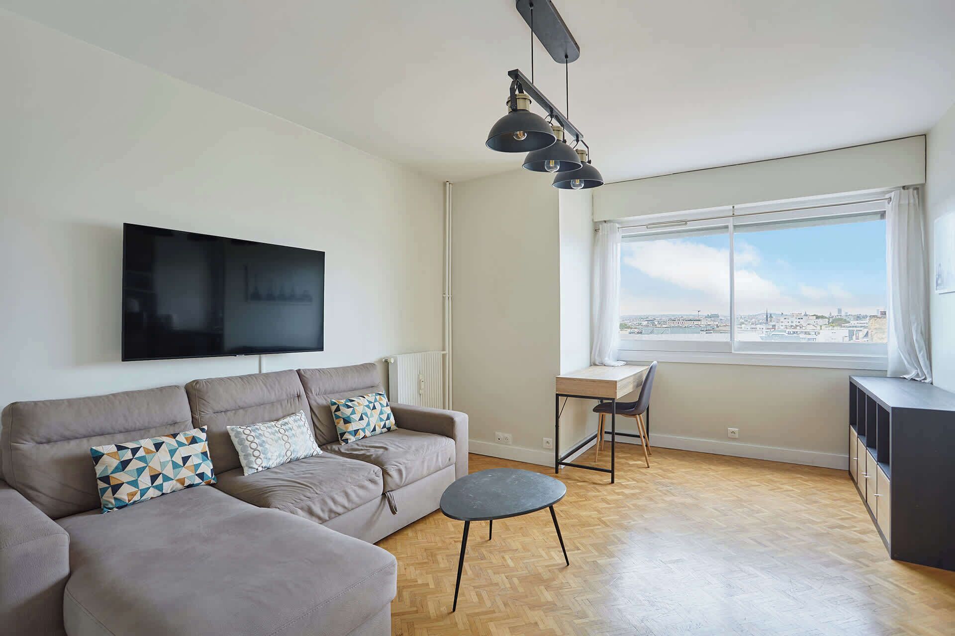 Ein modernes Wohnzimmer mit einer Couch, einem kleinen Tisch, einem an der Wand befestigten Fernseher und einem Fenster mit Blick auf die Stadt.