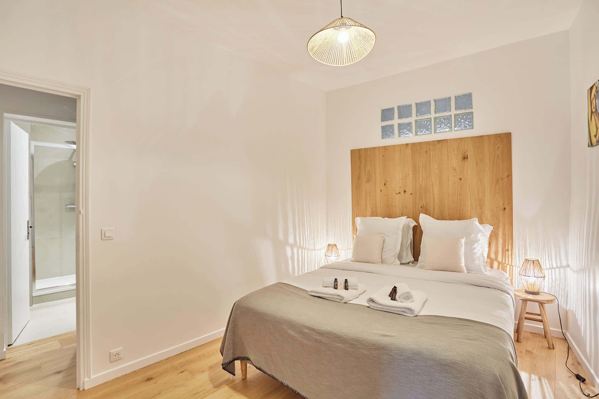 A bedroom with a wooden headboard, a bed with white linens, a bedside table with a lamp, and a door leading to a bathroom.