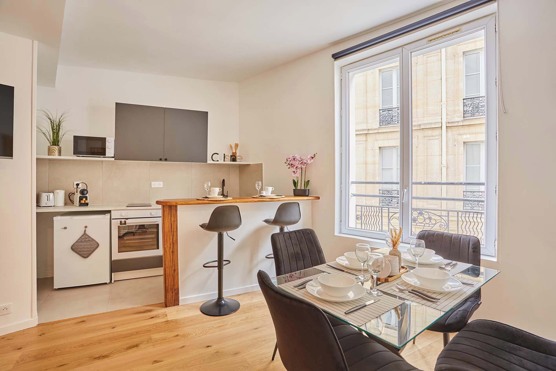 A compact apartment with a kitchenette, a dining area with a glass table, and a large window with a view of the city.