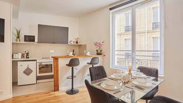 A compact apartment with a kitchenette, a dining area with a glass table, and a large window with a view of the city.