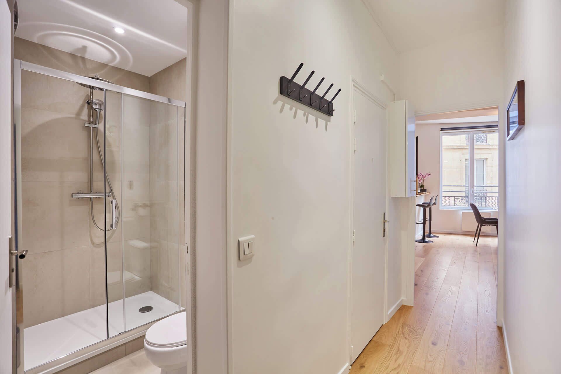 A modern bathroom with a glass-enclosed shower, a white toilet, and a hallway leading to a dining area with a view of the city.