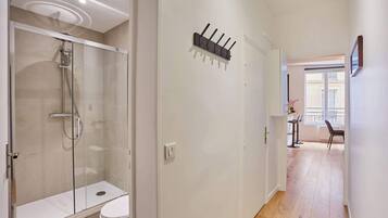A modern bathroom with a glass-enclosed shower, a white toilet, and a hallway leading to a dining area with a view of the city.