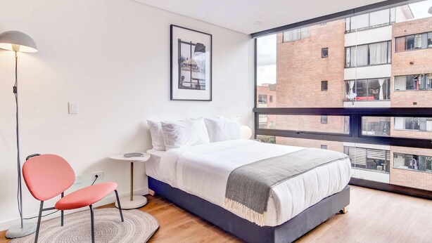 Une chambre moderne avec un grand lit, un fauteuil rouge, une lampe sur pied et une vue sur un bâtiment en briques, visible à travers une grande fenêtre.