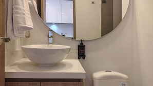 Un baño con un espejo ovalado grande, un lavamanos blanco, una toalla colgada en la pared y un inodoro con dispensador de gel antibacterial.