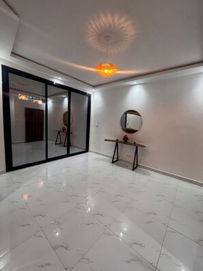 A spacious room with a large sliding glass door, a round mirror on a wooden stand, and a modern ceiling light fixture. - Atypical & Modern Duplex (Dakar)